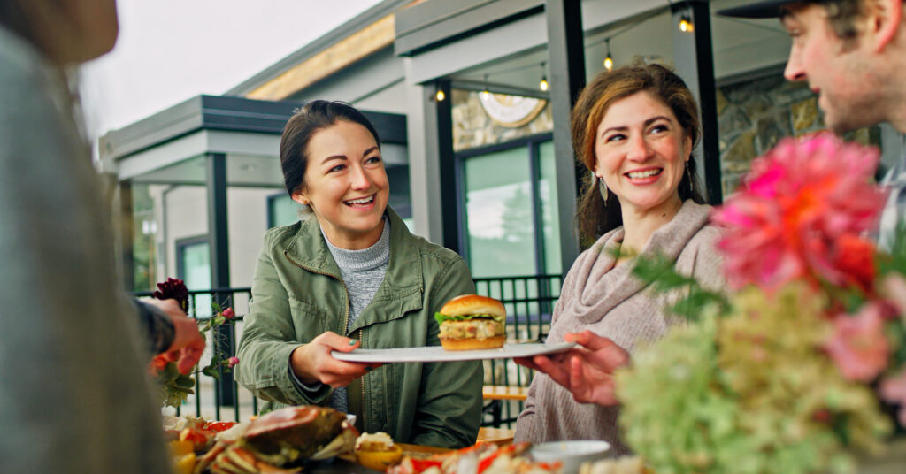 A group enjoying dinner on a patio, one woman is passing a plate with a fish burger to a man.