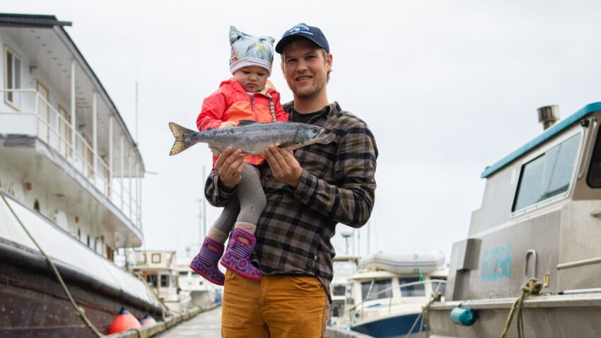A man holds a baby and a fish on a fishing boat dock.