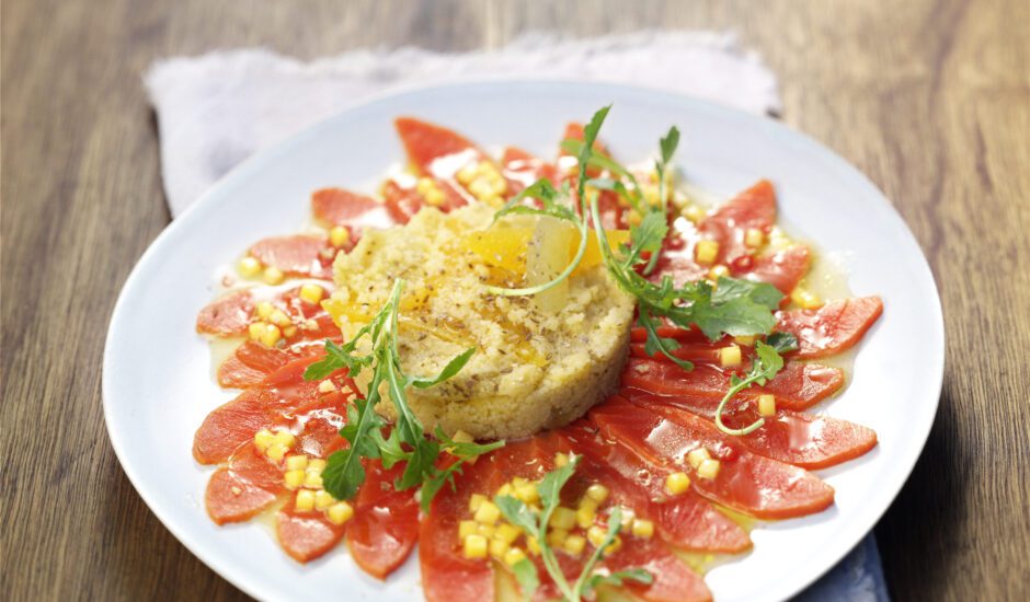 cured slices of salmon in a circular fanned pattern with couscous piled in center on top of a white plate with tan napkin and brown wood table