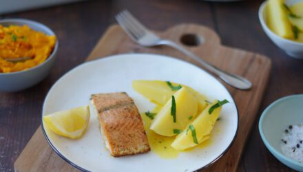 On a white plate lies a fillet of Wild Alaskan salmon with a lemon wedge and three potatoes garnished with herbs.
