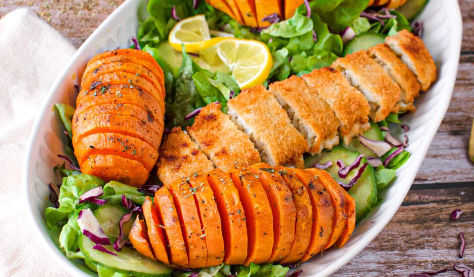 sliced breaded fish on a plate of green salad surrounded by sweet potatoes sliced in hasselback style