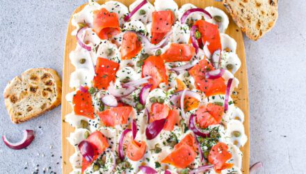 Plated on light wood cutting board is spread of cream cheese generously covered in slices of Alaska smoked salmon, capers, slices of red onion, dill, and sesame seeds. Slices of toasted bread lean in, waiting to be spread.