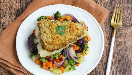 Plated on a colorful bed of red onion, potatoes, carrots, and broccoli, lays a fillet of Wild Alaskan Pacific cod crusted in a crunchy olive skin. The meal is garnished with a single leaf of parsley.