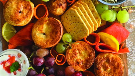 Lavish charcuterie board featuring many grapes, lemon slices, crackers, pretzels, artichoke hearts, tomatoes, and delicate ribbons of Alaska smoked salmon. These all pair beautifully with the wild Alaska salmon mini quiche featured in the middle of the image.