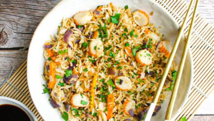 White ceramic bowl holds a lavish rice bowl with carrot, surimi, red onion, and herbs. A pair of chopsticks rests on the bowl.