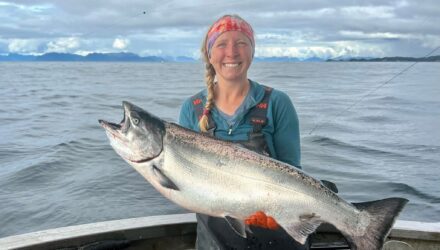 A woman with a blonde braid and pink headband shows off a king salmon on board a boat with ocean in the background.