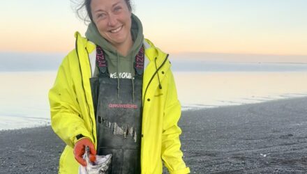 Fisher woman wearing a yellow jacket poses with a king salmon in one hand and holding a white fluffy dog's leash in the other.