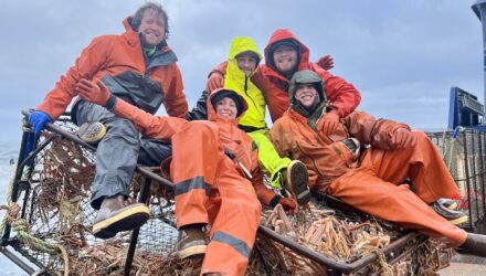Several people in orange rain gear jump on top of a truck bed filled with crab pots in celebration of a successful harvest.