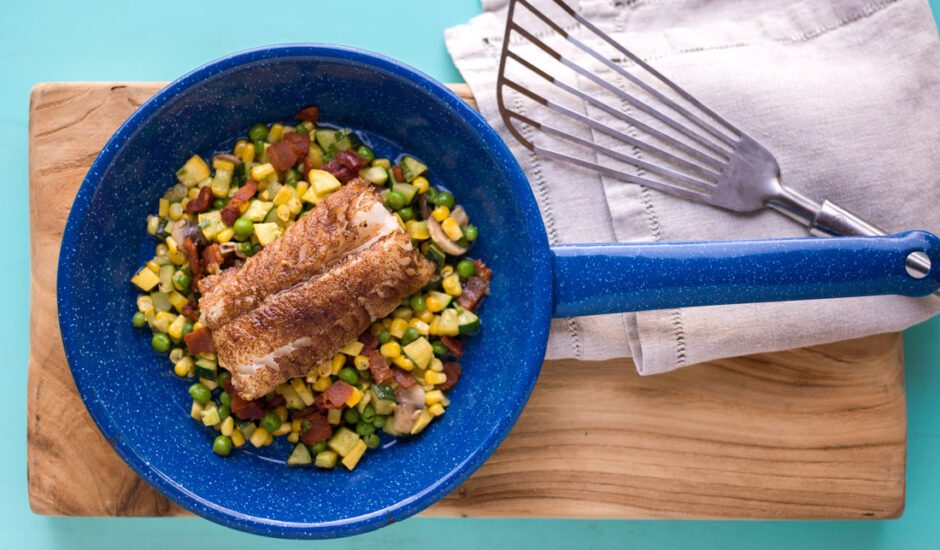 Browned fish on top of corn inside of vibrant blue pan on wood and teal background