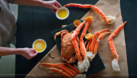 king crab and snow crab legs and a whole cooked dungeness crab sit on a brown triangle shaped piece of paper on a black table with hands bringing melted butter into the frame
